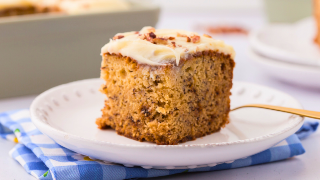 A slice of banana cake with cream cheese frosting on a plate. Perfect for snacking or serving as a dessert. Serve it with or without cream cheese frosting. DearCreatives.com