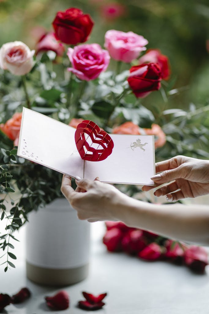 table with a lady's hands making a floral display in a vase and adding a heart shaped pick with card