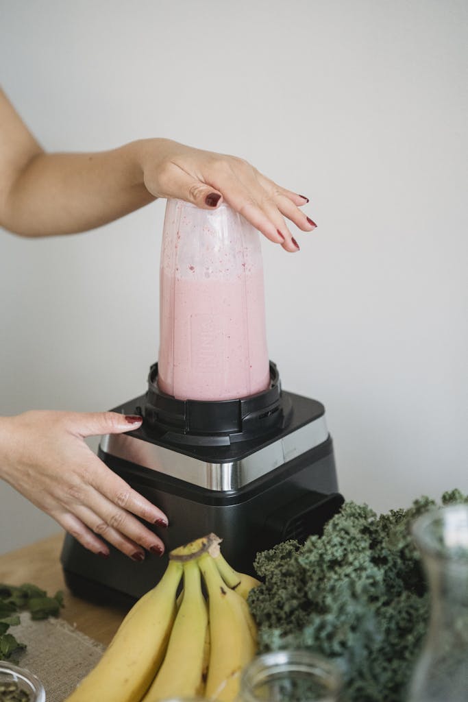 Hands making a nutritious pink smoothie with bananas and kale using a blender.